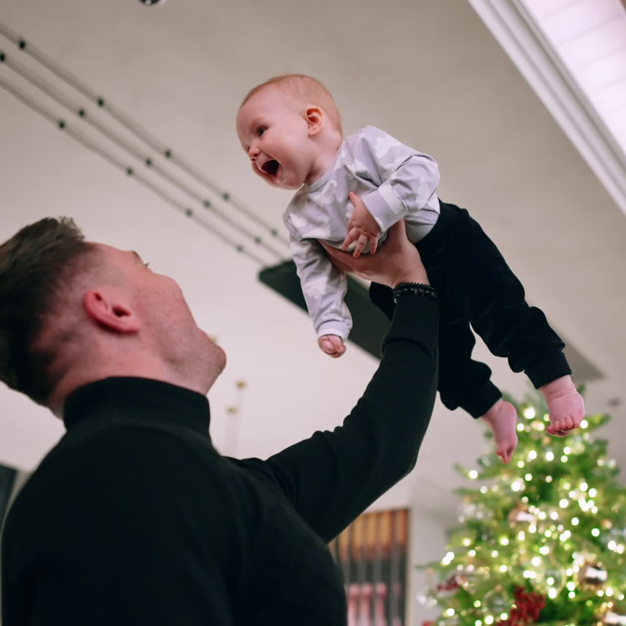 Man in black sweater holding his son over the head. Little child is excited and touches dad's face. Low angle view