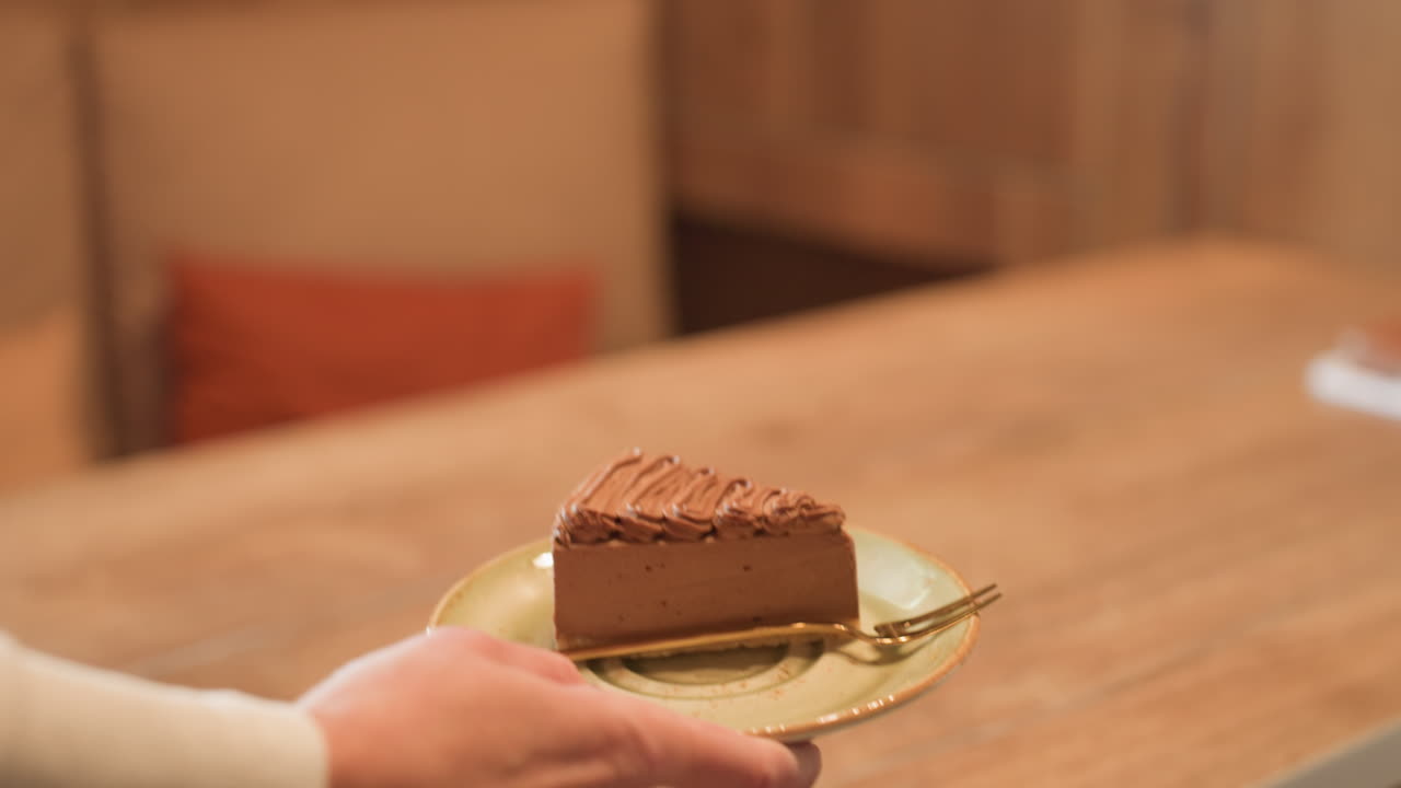 Close up of hand holding saucer with slice of chocolate cake and golden spoon, approaching table in cozy cafe setting with blurred background and soft warm lighting, notebook slightly visible