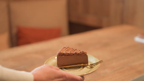 close up of hand holding saucer with slice of chocolate cake and golden spoon, approaching table in cozy cafe setting with blurred background and soft warm lighting, notebook slightly visible