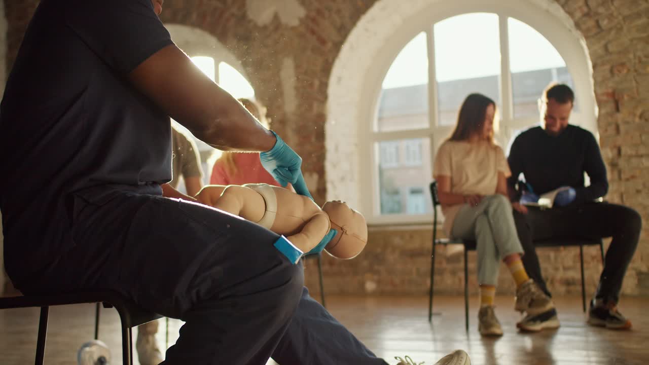 Close-up shooting: A Black male doctor in a blue medical uniform demonstrates the technique of artificial respiration of a child using the example of an infant mannequin for people studying. Course of a medic