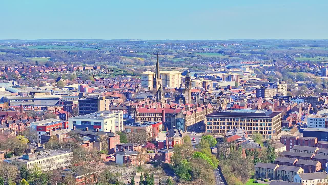 Aerial view approaching the Cathedral of All Saints in Wakefield, Yorkshire, England, with busy roads, surrounded by greenery, urban buildings, and a clear horizon stretching into the distance.