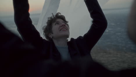 smiling woman holding translucent fabric flowing in breeze on frozen lake shore