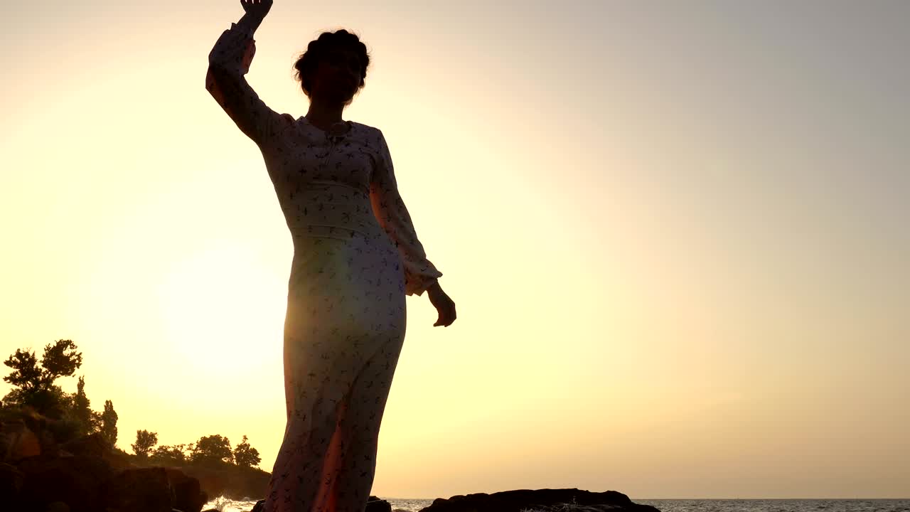 adorable chica caucásica morena en vestido claro bailando en el acantilado sobre el mar a la hora del amanecer