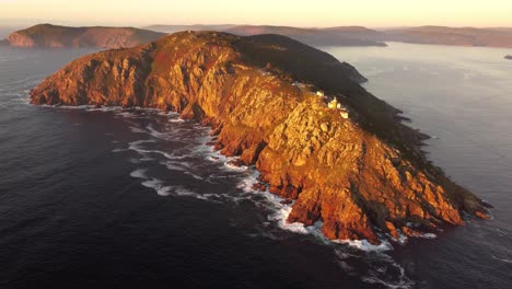 Vista-Aérea-Frontal-Del-Extremo-Europeo-De-La-Tierra-Cabo-Finisterre-Cabo-Fisterra-Durante-La-Puesta-De-Sol,-Capilla-Del-Faro-En-La-Cima-De-La-Formación-Montañosa-De-Acantilados-Rocosos-En-El-Océano-Atlántico