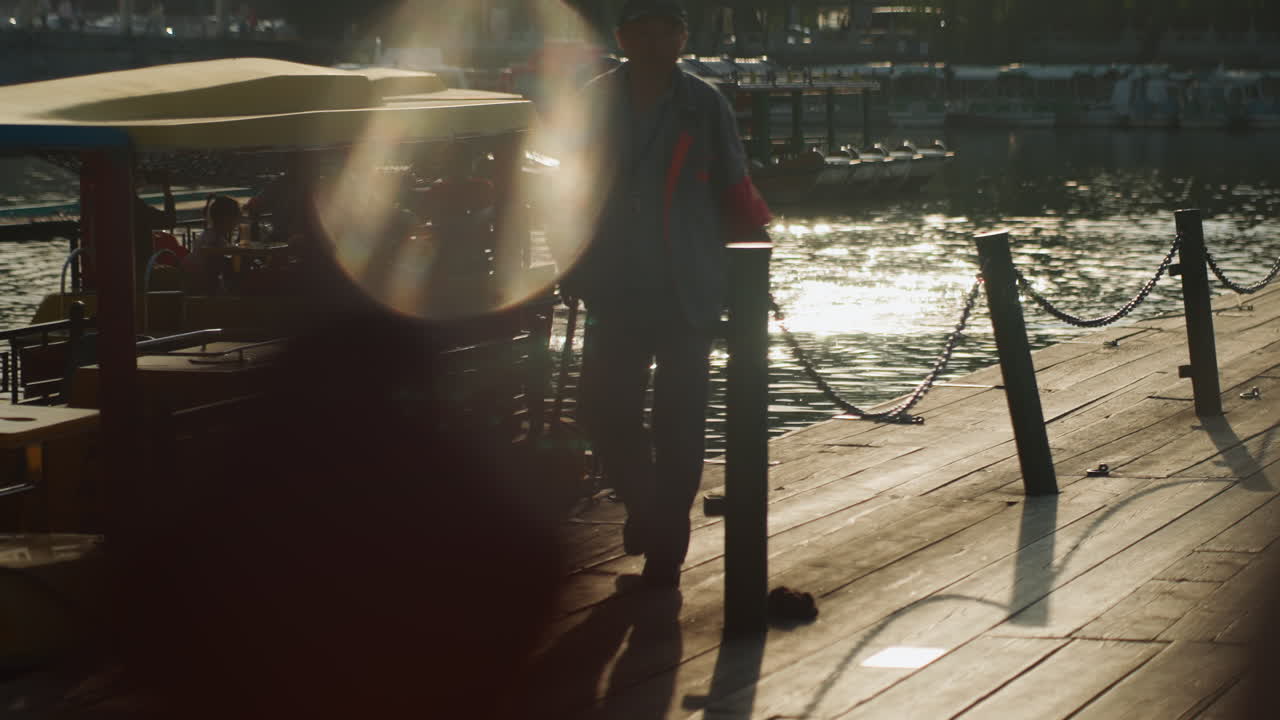 Mann geht auf sonnenbeschienenem Steg zur goldenen Stunde, hintergrundbeleuchtete Silhouette überquert Uferpromenade nahe festgemachtem Boot und Kiosk, starkes Lens Flare und schimmernde Reflexionen auf sich kräuselndem Wasser, stimmungsvolle cineastische Atmosphäre