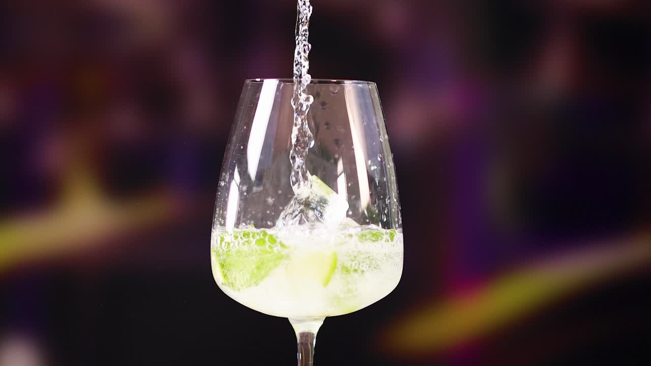 A mojito cocktail is poured into a glass with lime and mint, set against a vibrant background