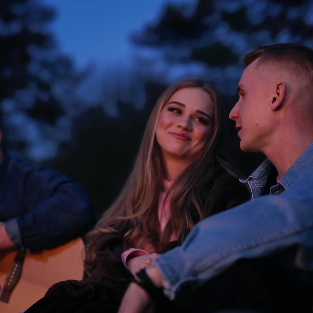 Young friends rest in nature at dusk. Beautiful woman listen to a boyfriend while sitting together at camping party. Guy playing the guitar