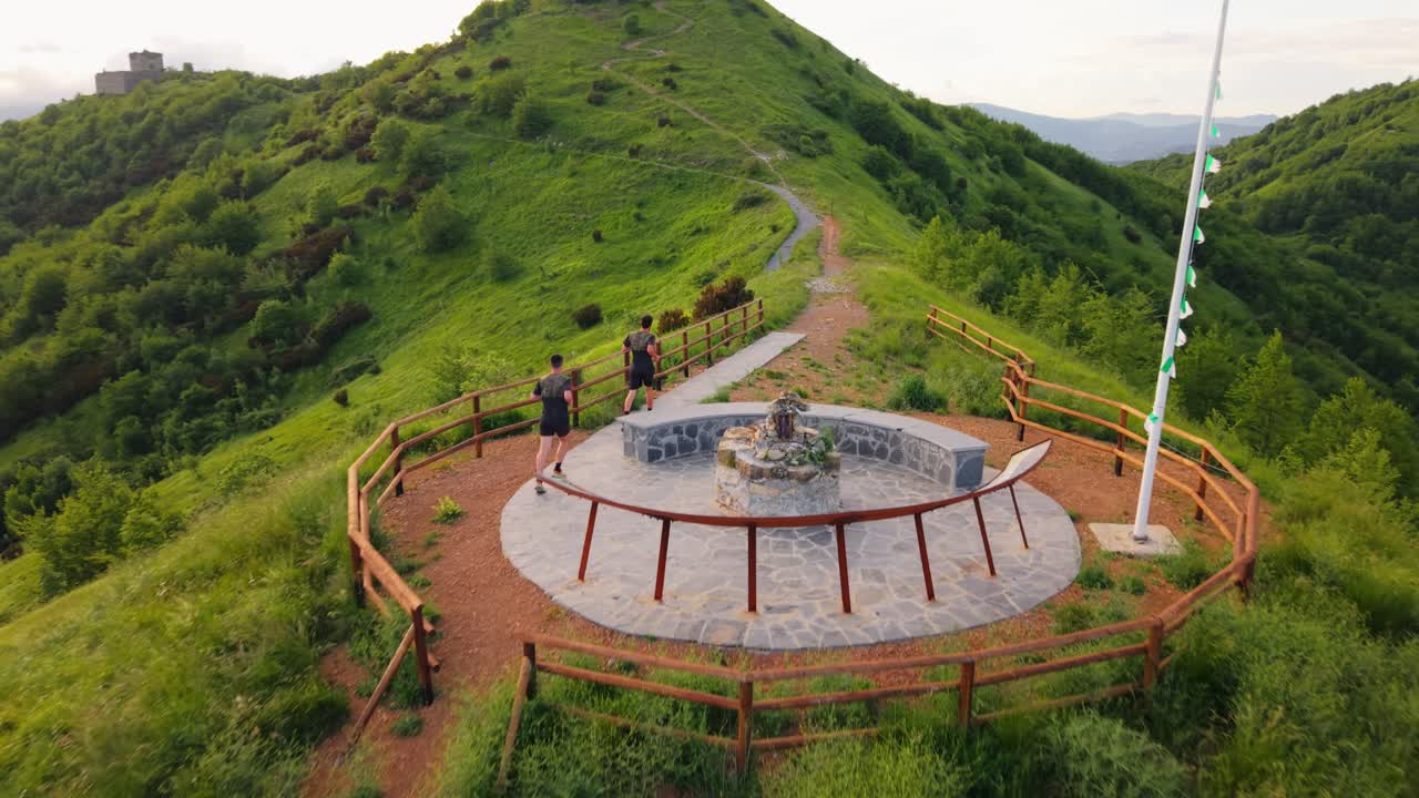 Two men running along a mountain trail surrounded by lush greenery and hills