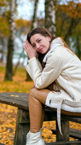 relaxed calm dark-haired lady sits on the table thinking of something. caucasian girl enjoying beautiful autumn day in the park among birch trees. vertical video