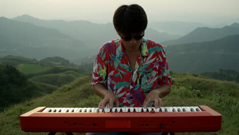 Un-Músico-Asiático-Toca-Un-Piano-Rojo-Al-Aire-Libre-En-Un-Paisaje-Natural,-Usa-Ropa-Vintage,-Camisa-Hawaiana-Y-Gafas-De-Sol