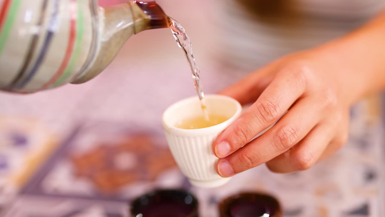 A person pours aromatic tea from a striped teapot into a small cup, captured in warm lighting