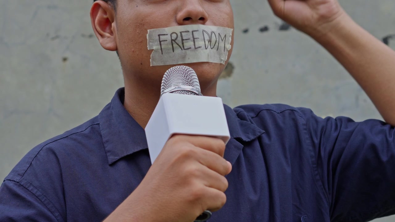 Close-up wartawan yang dibungkam dengan lakban bertuliskan "FREEDOM" di mulutnya, memegang mikrofon dan mengangkat kepalan tangan