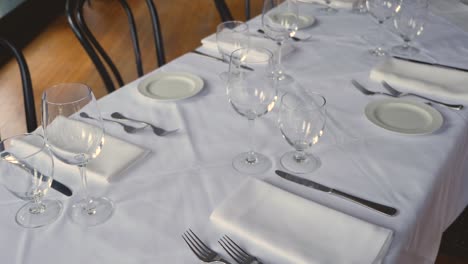 Dining table set for a wedding or corporate event at fine dining restaurant ceramic plates forks knives cloth napkins on white tablecloth on table steady slow motion medium-wide panning down left