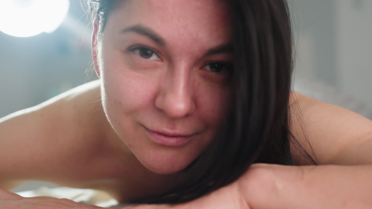 Beautiful model lifts head with soft smile while resting face down bare back on spa massage bed under warm natural light creating tranquil blissful atmosphere of relaxation and comfort