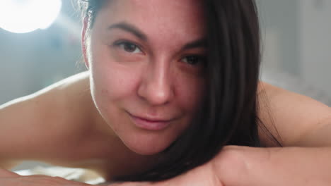 beautiful model lifts head with soft smile while resting face down bare back on spa massage bed under warm natural light creating tranquil blissful atmosphere of relaxation and comfort