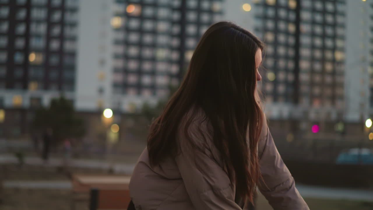 A girl in a peach jacket and black trousers is seen skating towards a bench in an urban park. After sitting down, she rests her hand on the bench, with her face turned slightly away from the camera