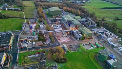 Slow-sweeping-aerial-dronefootage-of-a-large-industrial-plant-at-dusk,-showing-pipework-structures,-buildings,-cooling-towers,-steam,-and-work-vehicles