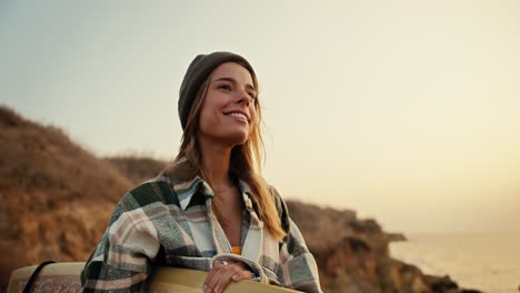 Close-up-shot-of-a-blonde-girl-in-a-green-hat-and-a-plaid-shirt-holding-a-yellow-surfboard-in-her-hands-standing-and-looking-at-the-sea-near-a-rocky-shore-by-the-sea-in-the-morning-at-sunrise