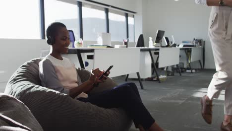 African-american-businesswoman-wearing-headphones,-sitting-in-armchair-while-her-coworker-passing-by