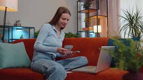 Smiling-happy-girl-counting-money-cash-and-use-laptop-pc-calculate-domestic-income-earnings-at-home