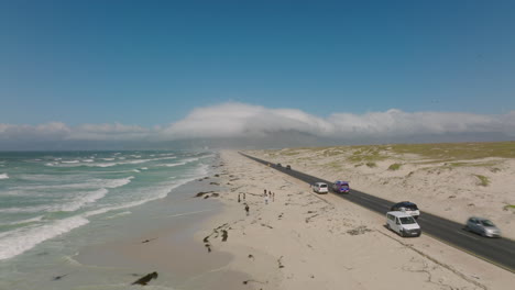 Cars-driving-on-road-along-coast.-People-taking-rest-and-walking-on-sandy-beach.-Waves-washing-shore.