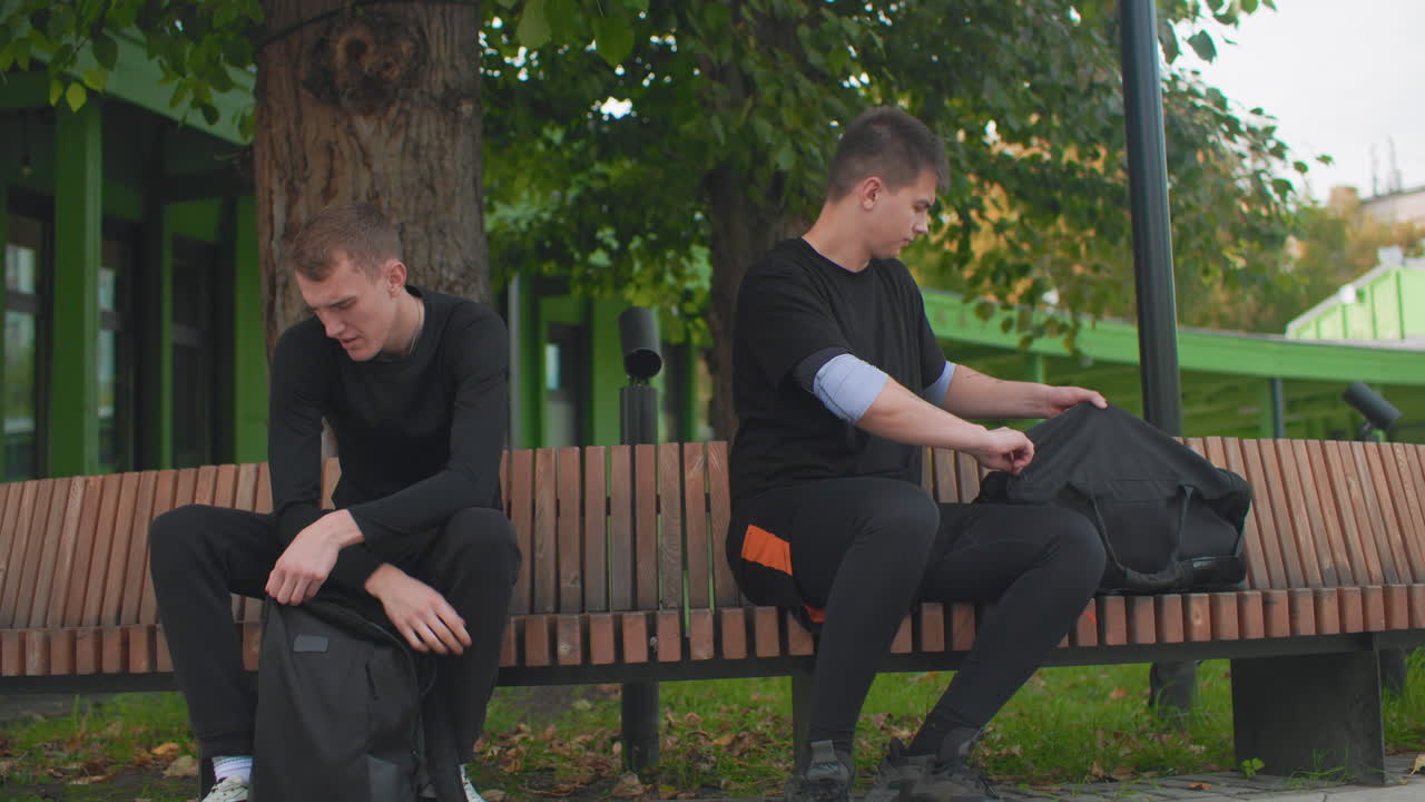 Two white men on park bench under tree, one opens backpack and checks shoulder tattoo while awkward glance establishes tension, green shelter and autumn leaves in background, casual black clothing