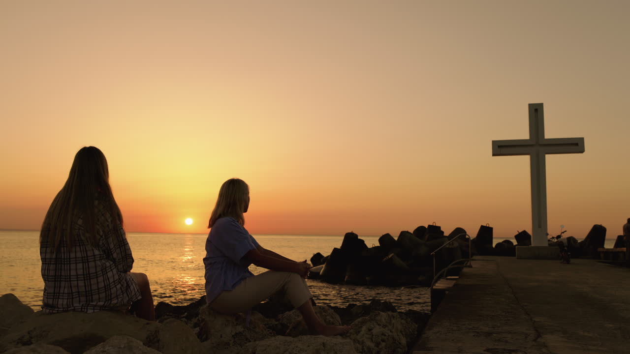 Two women watching the sunset by the sea with a cross