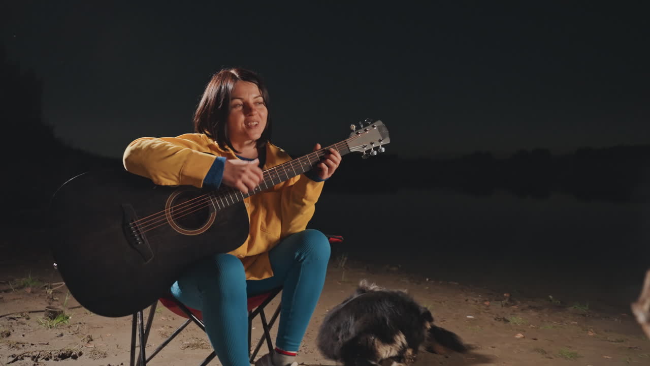 Ung kvinde spiller guitar ved et lejrbål på en sandstrand om natten, gul jakke, klapstol, blød røg og varm glød, blid strumming og intim melodi, eftertænksom stemning og rolig atmosfære.