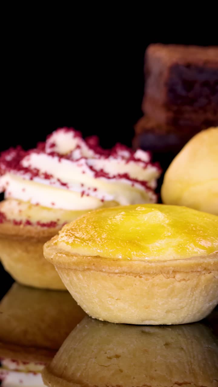 Una lenta carrellata della telecamera rivela brownies, cupcake, crostate e fragole fresche disposti su uno sfondo nero riflettente sotto un'illuminazione brillante e uniforme.