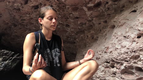 tilt finds blond girl meditating inside cave, with rocky background