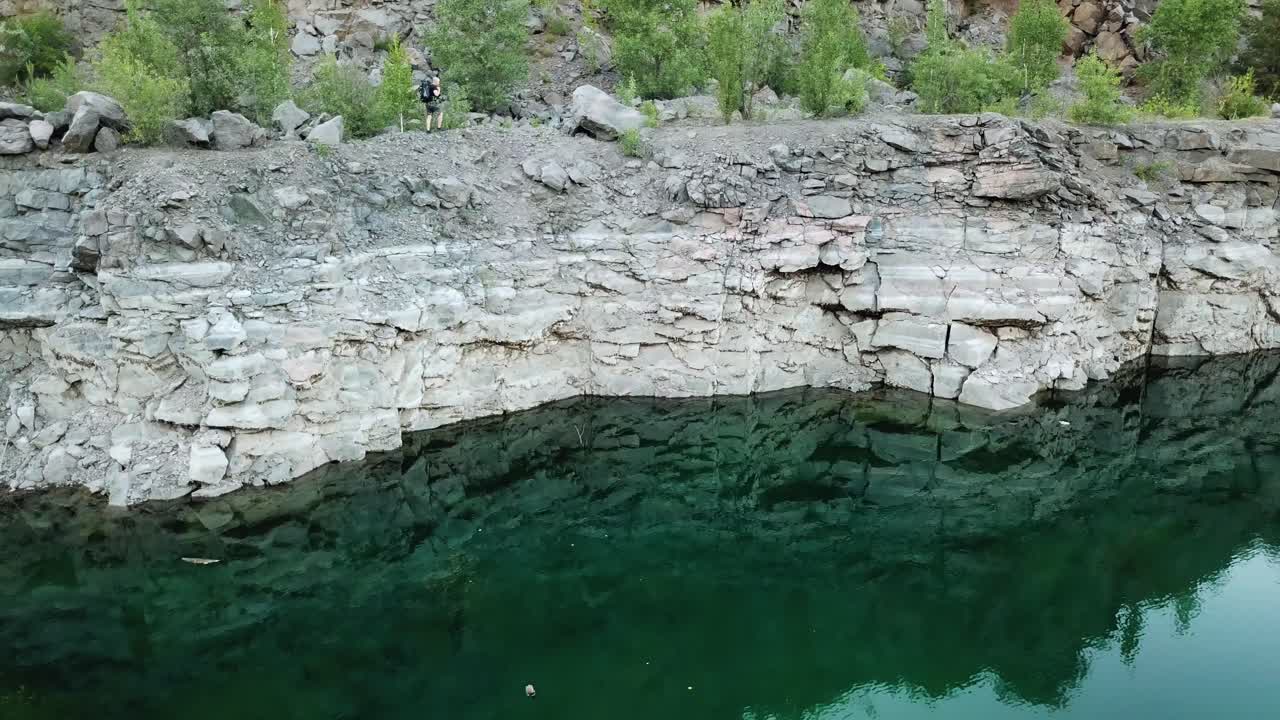 Aerial view of a young traveler with a backpack walking along a rocky path. Rocky shoreline of the lake with forest. Camping season