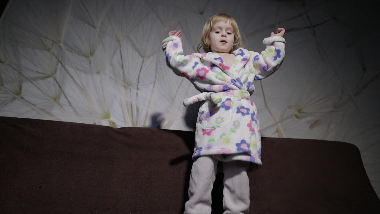 Little cute girl with blond hair jumps on sofa. Bathrobe clothes. Happy