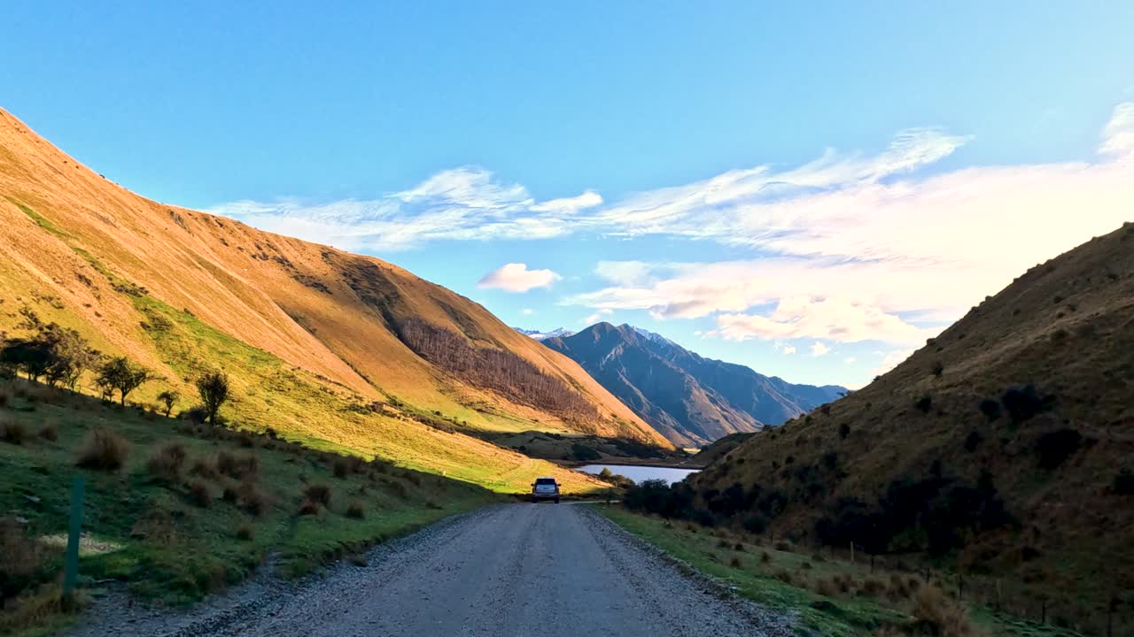 Vehicle travels scenic gravel road between hills toward lake, mountains, under bright daylight, steady camera