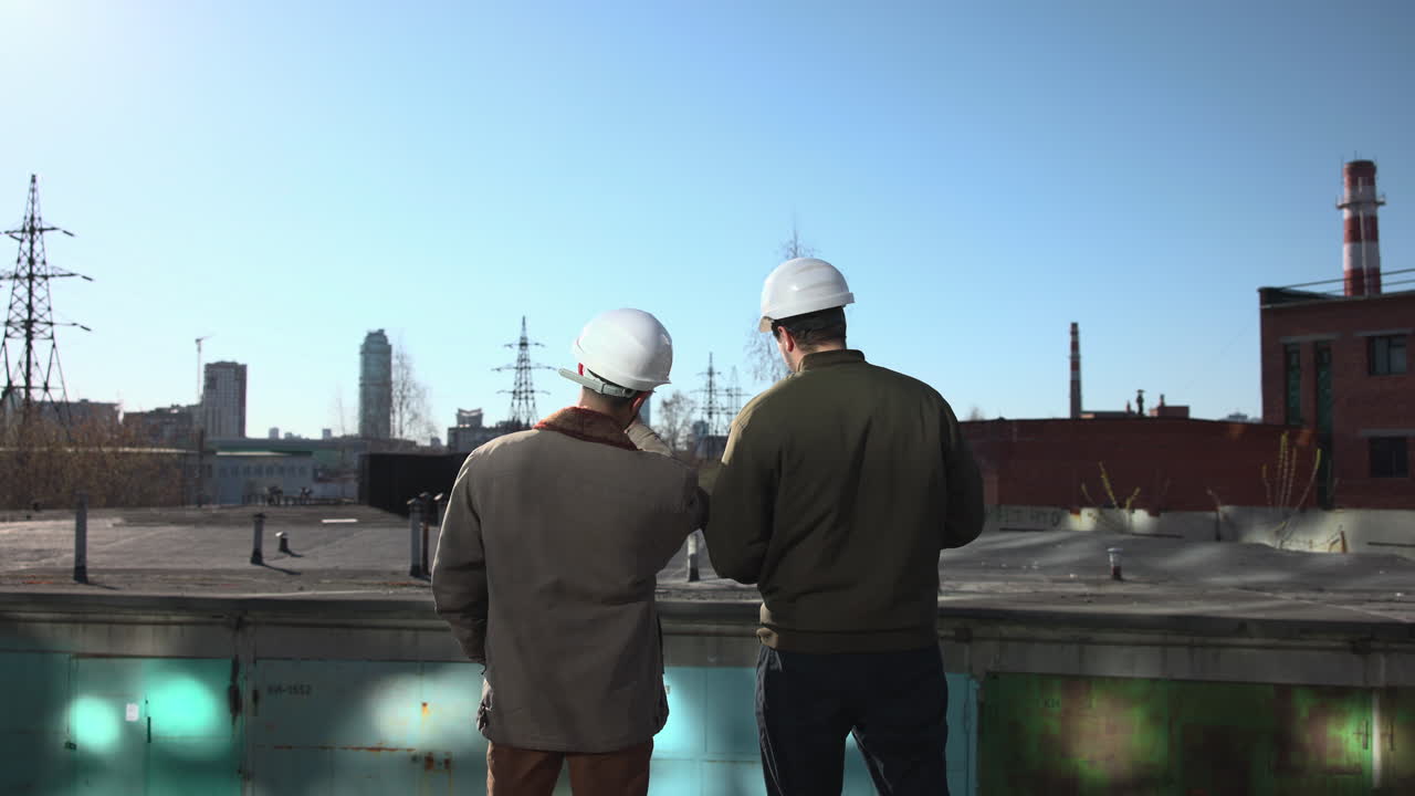 Two Caucasian Engineers On Rooftop Inspecting Plans, Pointing Toward Industrial Skyline, Wearing White Hard