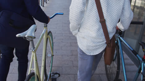 Two-diverse-male-friends-walking-together-wheeling-bicycles-in-street