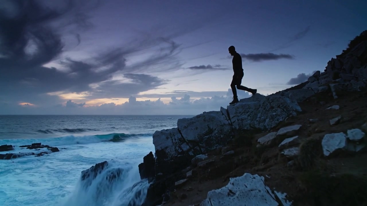 Uomo con giacca e stivali che scende da una scogliera rocciosa al tramonto, dopo che un'onda ha spruzzato acqua, mentre osserva, con spazio per il testo.