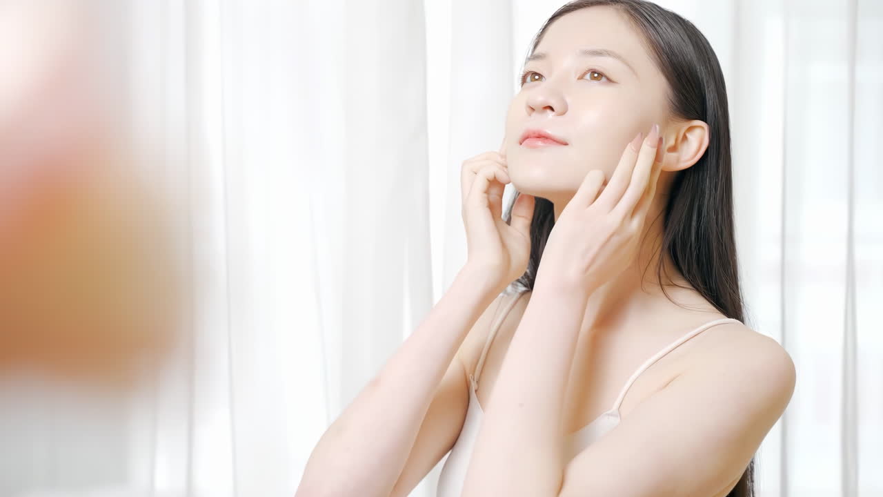 美しい若い女性が、顔にクリームを塗った後、指で肌の弾力と潤いをチェックしている。保湿化粧品によるスキンケアや皮膚科治療に関心があり、自宅でハンドクリームを楽しむ女性。