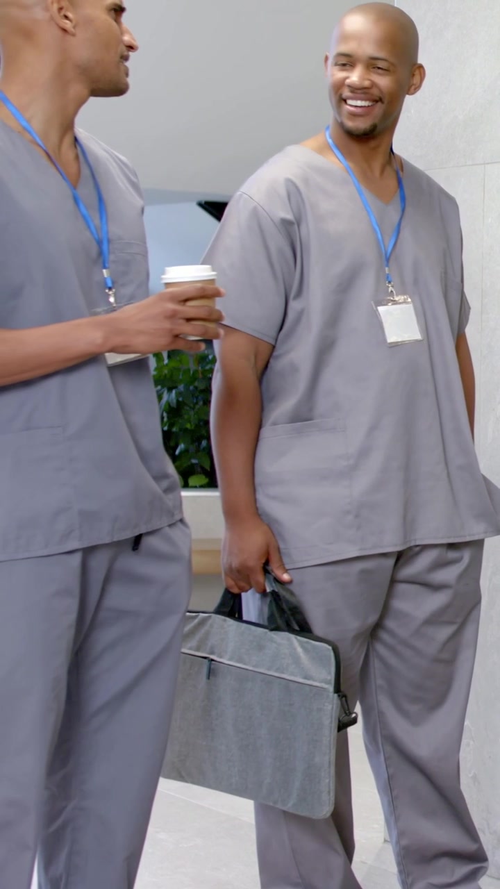 Video vertikal: Dua staf berjalan setelah shift di lobi dengan seragam medis, memegang kopi, tas laptop, mengobrol.