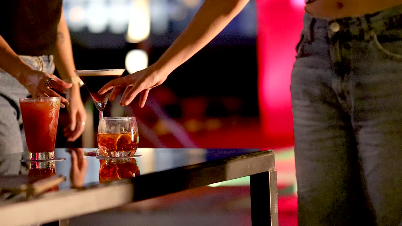 Two women in casual attire select cocktails from a table in vibrant rooftop bar lighting