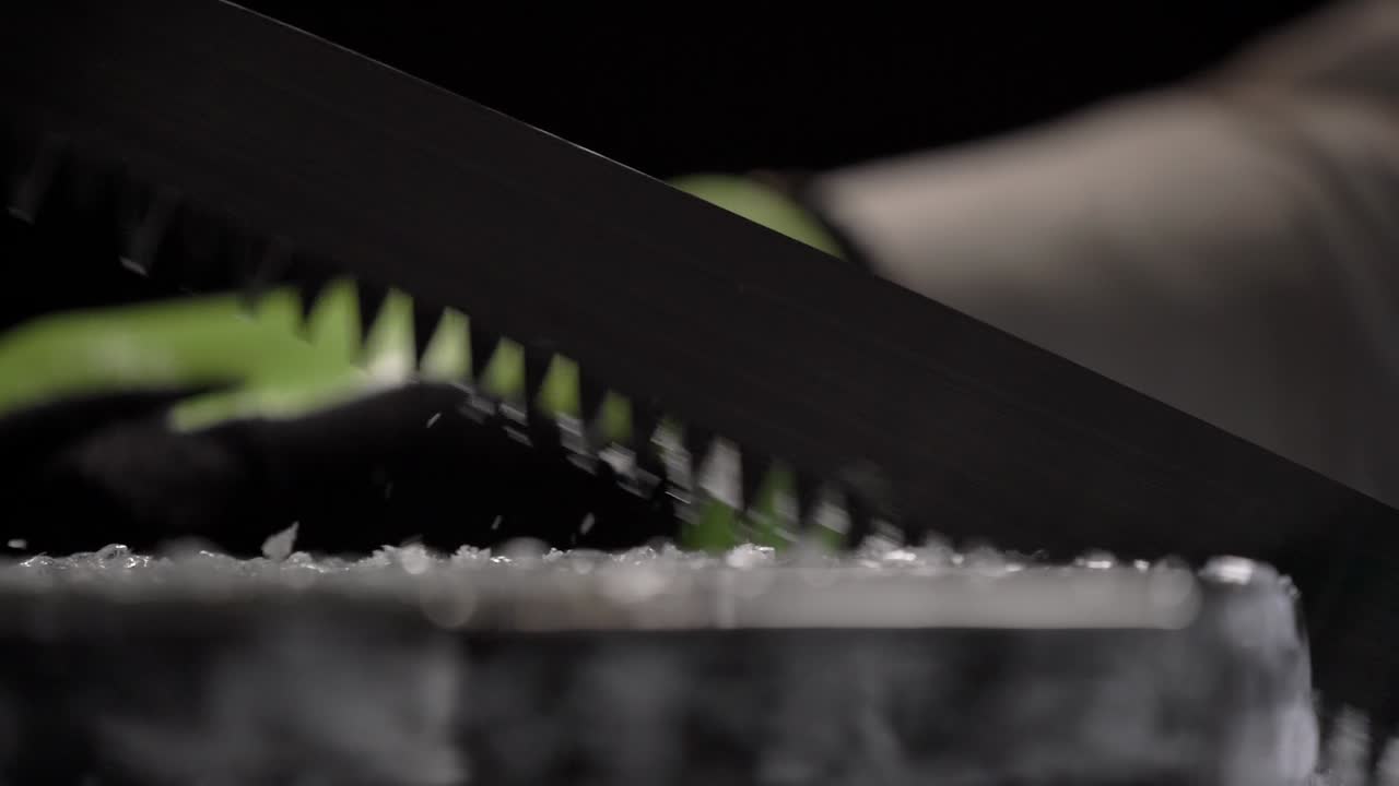 A bartender is cutting the huge perfectly transparent ice block using a long toothed knife and protecting gloves.