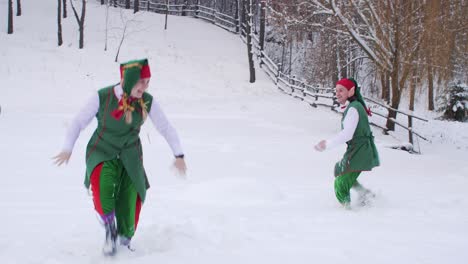 two cheerful elves in green clothes play snowballs near the winter forest
