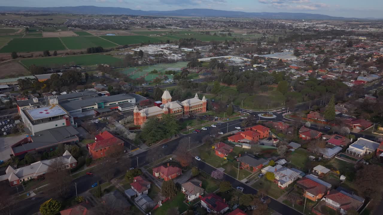 Aerial drone pullback over urban neighborhood toward hospital with soft warm evening light and trees, Bathurst NSW Australia