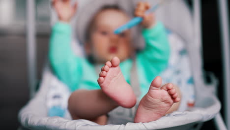 tiny feet of an infant sitting in a baby chair. cute child pulls spoon into mouth. blurred backdrop.