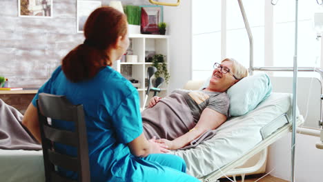 in bright retirement home old lady lying in hospital bed talks with a female caregiver