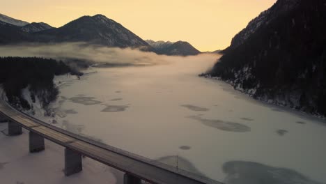 Sylvenstein-fresh-drinking-water-reservoir-bridge-in-the-beautiful-Bavarian-Austrian-alps-in-snow-winter-with-mountains-and-clear-blue-water-by-sunset