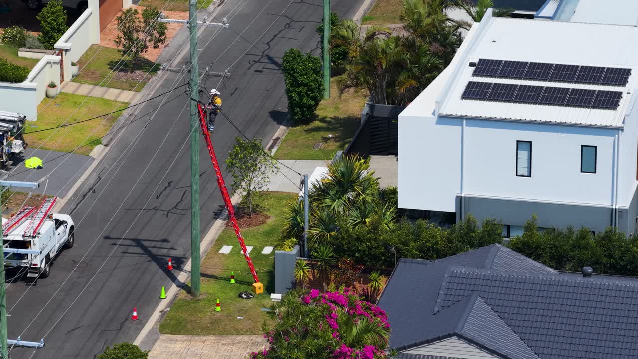 Trabalhador de serviços de utilidade pública de alta visibilidade em poste de energia a partir de escada em rua suburbana australiana ensolarada, visão aérea