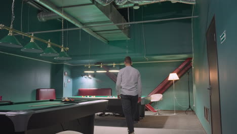 back shot of player in white shirt and grey trousers walking into billiard room, approaching pool table, and picking up cue stick. overhead lamps illuminate green pool tables