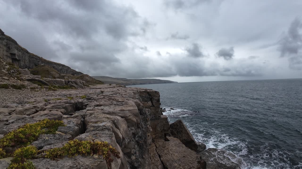 Drone footage captures dramatic waves crashing against rugged ledge rock formations in Dorset's Isle of Purbeck. Moody skies and powerful sea evoke nature's raw power