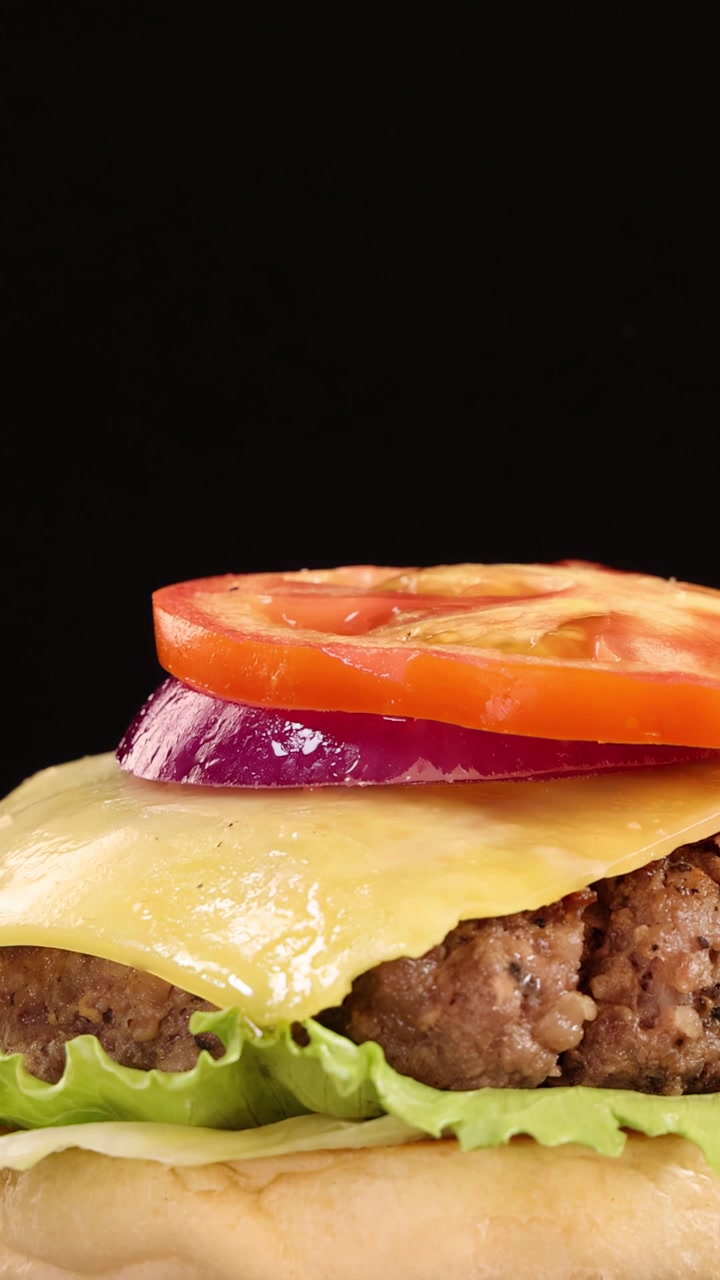 La parte superiore di un panino ai semi di sesamo cade al rallentatore su un cheeseburger di manzo impilato con lattuga, cheddar, pomodoro e cipolla rossa, ripreso su uno sfondo nero con un'illuminazione brillante e uniforme