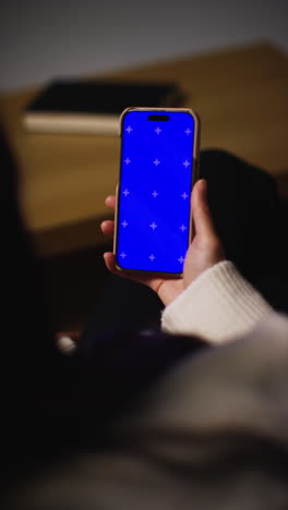 vertical video over the shoulder shot of woman at home sitting on sofa with blue screen mobile phone scrolling through internet or social media